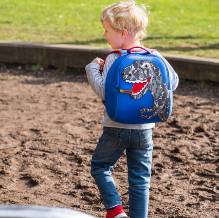 Young blonde boy wearing a blue and red Dabbawalla T-Rex dinosaur backpack and walking in the sand.