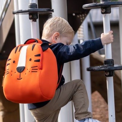 Dabbawalla Bags Tiger backpack featuring smiling face of tiger with black and white applique on orange body.Active young blonde boy climbing play structure with  bright orange and black tiger backpack.