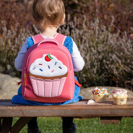 Small girl sitting on a bench wearing a pink backpack.  Backpack features a large cupcake applique on front panel. 
