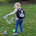 Preschool girl wearing a French Bulldog Dabbawalla Backpack and making bubbles is the grass.