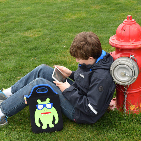 Young boy leaning against a hydrant engrossed in his tablet.  The monster geek tablet case by Dabbawlla Bags is resting against his leg.